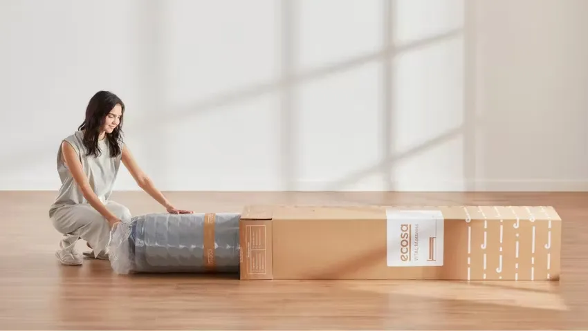 Ecosa Mattress Packaging A woman kneeling next to a rolled-up Ecosa mattress being unpacked from a branded cardboard box.